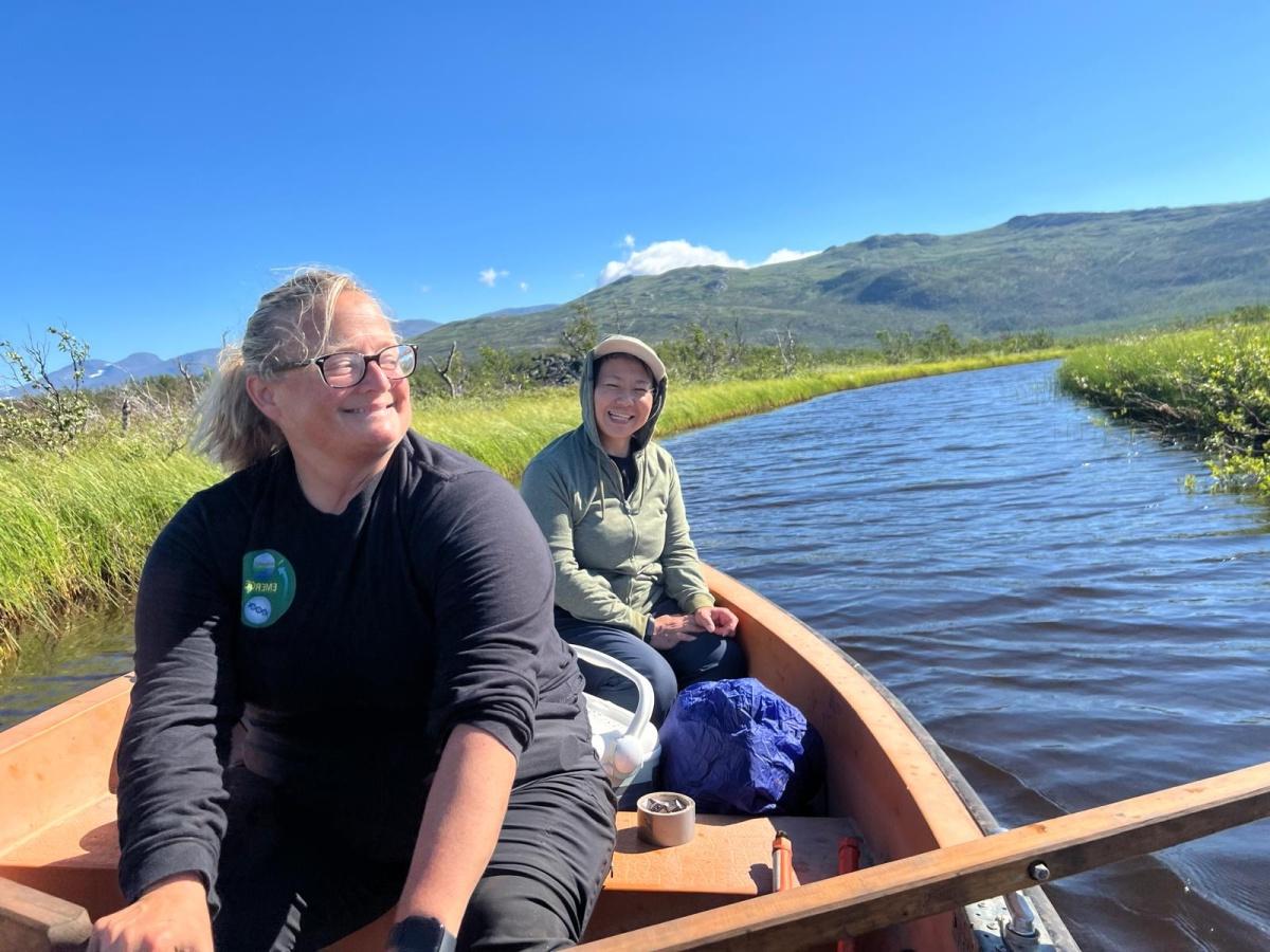 Two women row a wooden boat beneath snow-capped mountains.