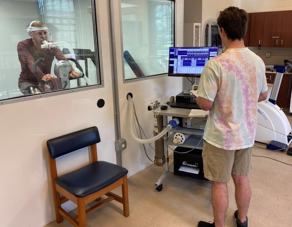 Researcher looks at a computer while a subject rides an exercise bicycle behind glass